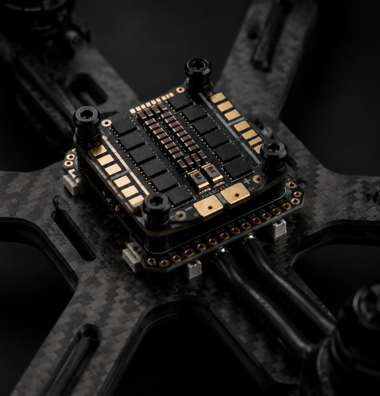 A close-up of a black electronic flight controller or drone processor mounted on a carbon fiber drone frame, with gold and silver metallic components and screws visible against a dark background.