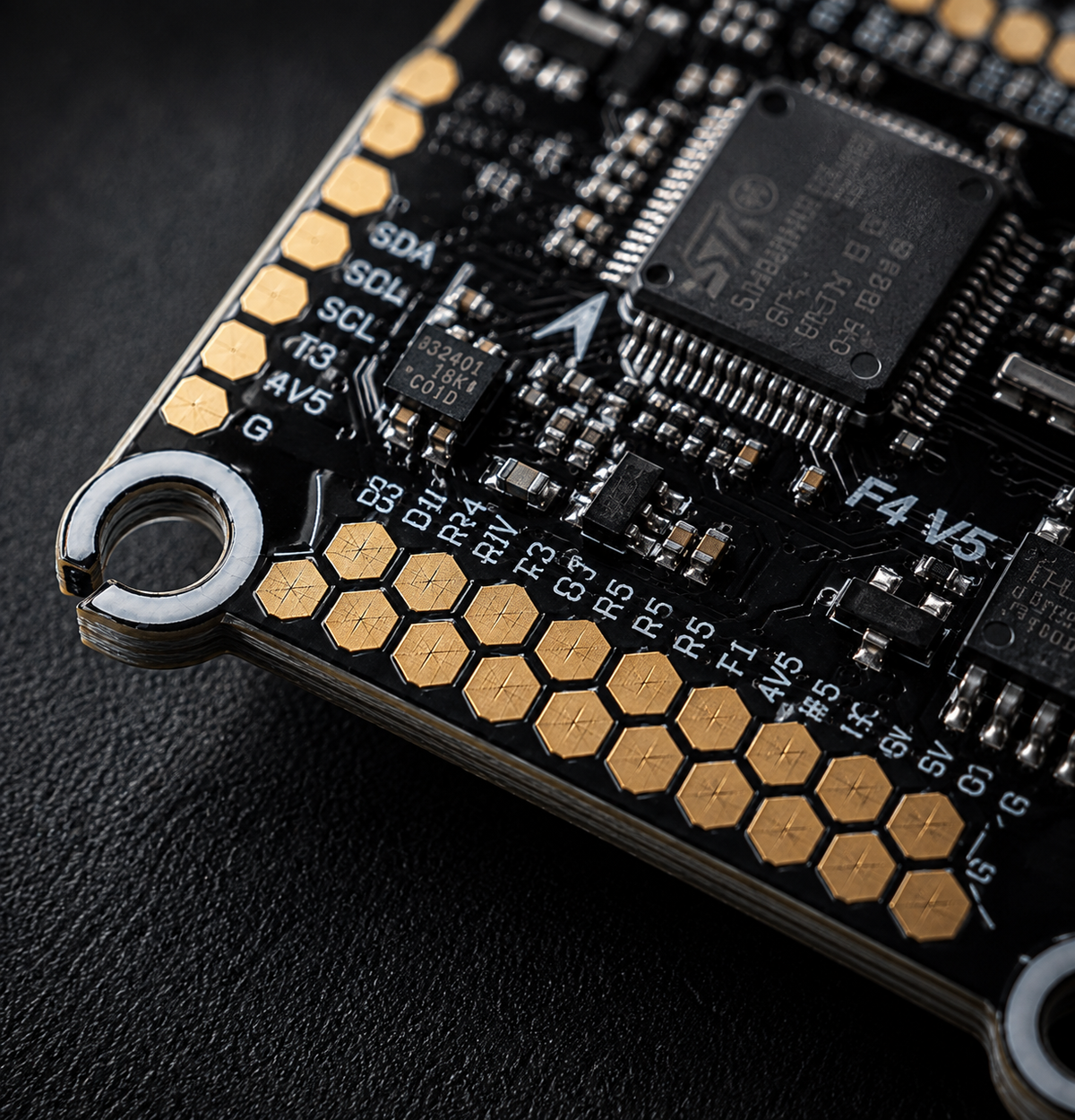 Close-up of a black electronic circuit board with gold hexagonal pads, a large central processor chip, and white printed labels on a dark textured background.